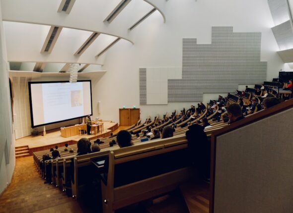 Sala de aula universitária moderna com alunos usando laptops e quadro digital ao fundo.